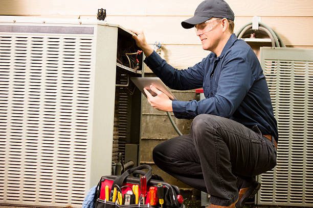 Repairmen works on a home's air conditioner unit outdoors. He is checking the compressor inside the unit using a digital tablet. He wears a navy blue uniform and his safety glasses. Tools inside toolbox on ground.